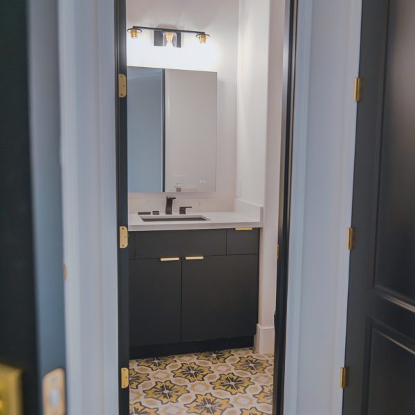 Modern Bathroom Vanity with Brass Accents A view through a dark doorway into a modern bathroom featuring a dark vanity with a white countertop and brass pulls, a rectangular mirror, and a vanity light above it, all set against a patterned floor.